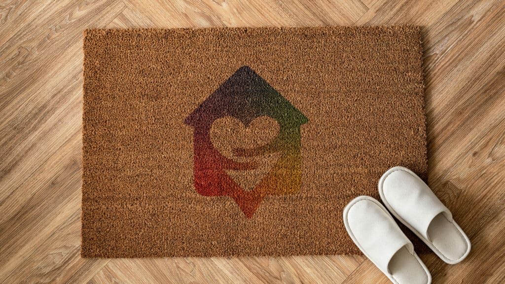 Doormat with a colourful Heartland logo featuring a heart and house design, placed on a wooden floor with white slippers beside it.